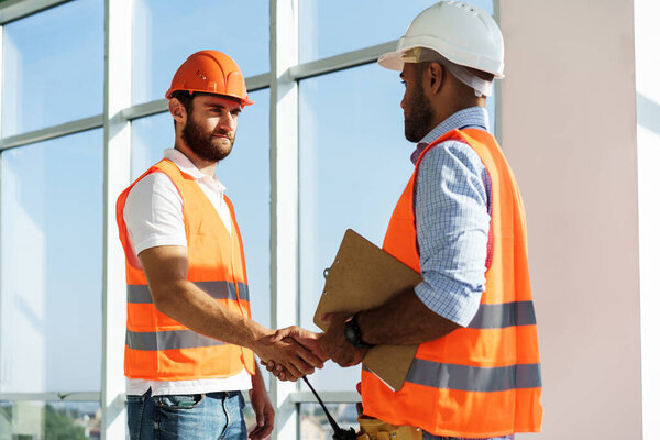Two men engineers in workwear shaking hands against construction site.