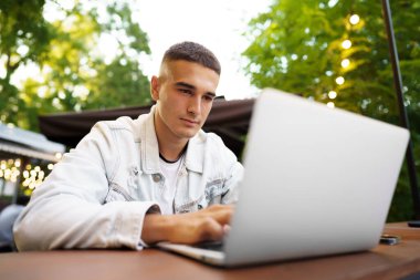 Genç adam dışarıda kafede çalışırken masada oturup dizüstü bilgisayarda klavye çalıyor.