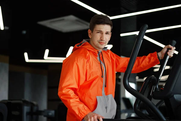 Portrét mladého muže v oranžové větrovce cvičení na fitness stroji v tělocvičně. — Stock fotografie