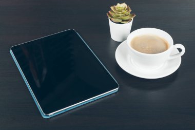 Siyah ekranlı dijital tablet, kahve fincanı ve çalışma masasındaki küçük bitki.