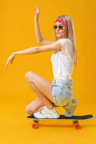 Skateborder Mädchen in kurzen Hosen und T-Shirt sitzt an Bord über gelbem Hintergrund — Stockfoto