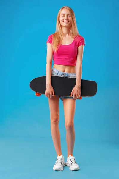 Attraktive junge Frau hält ihr Skateboard über blauem Hintergrund — Stockfoto