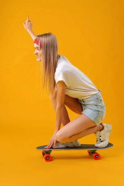 Korcsolyás lány rövidnadrágban és pólóban ül a fedélzeten sárga háttér felett — Stock Fotó