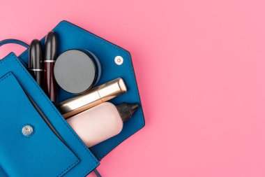 Parlak pembe arka planda kozmetik ürünlerle dolu küçük bir kadın çantası.