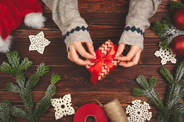 Manos en suéter sosteniendo un regalo en mesa de madera con decoraciones navideñas — Foto de Stock