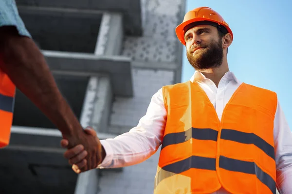 Två män ingenjörer i arbetskläder skakar hand mot byggarbetsplatsen. — Stockfoto