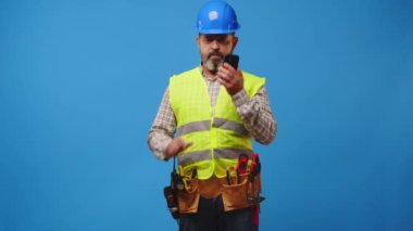 Mavi arka planda akıllı telefonunu kullanarak yelek ve şapka giyen kıdemli inşaatçı.