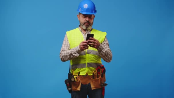 Senior man builder in vest en hardhat met zijn smartphone tegen een blauwe achtergrond — Stockvideo