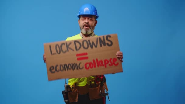 Senior man builder in hardhat holding cardboard placard striking against blue background — Stock video