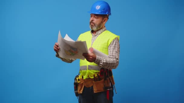 Senior man bouwer in hardhat op zoek naar blauwdrukken tegen een blauwe achtergrond — Stockvideo