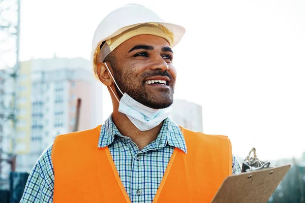 Porträtt av blandras man byggare i arbetskläder och hårdhatt bär medicinsk mask, närbild — Stockfoto