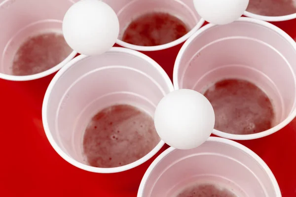 Poharak és műanyag labda piros háttérrel. Sörpong játék — Stock Fotó
