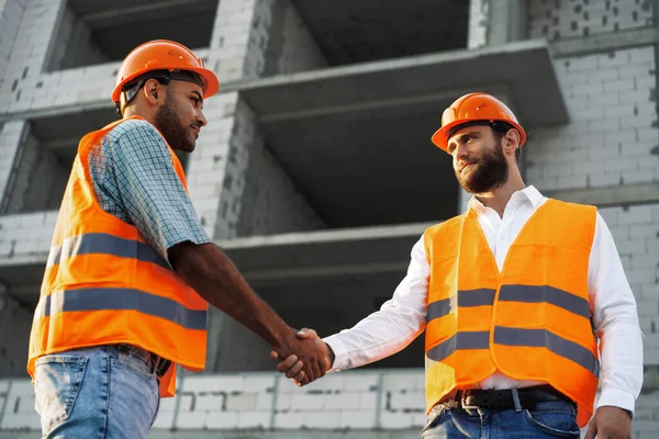 Två män ingenjörer i arbetskläder skakar hand mot byggarbetsplatsen. — Stockfoto