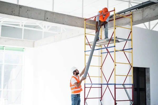 Üniformalı iki adam inşaat alanındaki iskelede boruyu sürüklüyor.