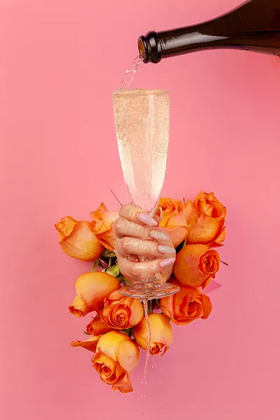 Mano femminile con manicure versando champagne in un bicchiere attraverso un buco in una carta strappata decorata con rose — Foto Stock