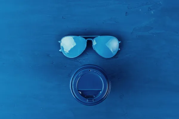 Vista superior de la taza de café para llevar con gafas de sol aviador sobre fondo azul clásico —  Fotos de Stock