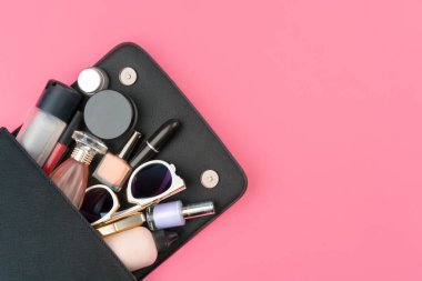 Parlak pembe arka planda kozmetik ürünlerle dolu küçük bir kadın çantası.
