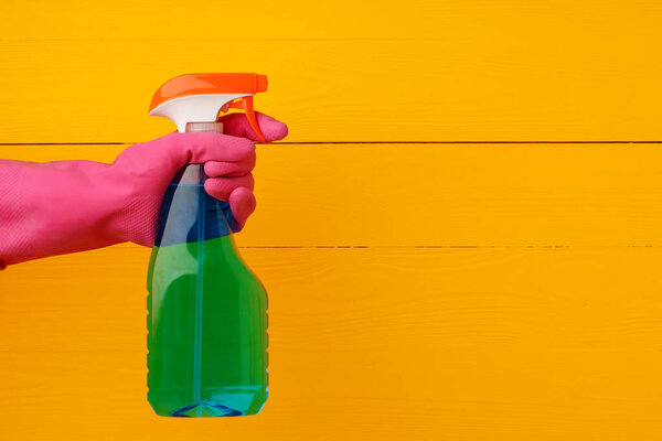 Перчатка, держащая в руках бытовой моющий спрей на желтом деревянном фоне