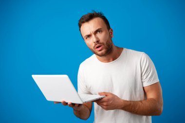 Yakışıklı genç adam dizüstü bilgisayarını mavi arka planda kullanıyor.