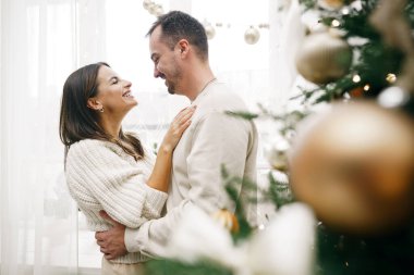 Genç çift evde Noel ağacının yanında sarılıyor.