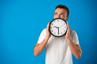 Beyaz tişörtlü yakışıklı genç adam mavi arka planda saatle poz veriyor.