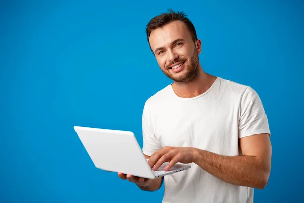Yakışıklı genç adam dizüstü bilgisayarını mavi arka planda kullanıyor. — Stok fotoğraf