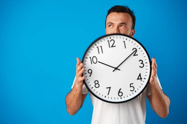 Beyaz tişörtlü yakışıklı genç adam mavi arka planda saatle poz veriyor.