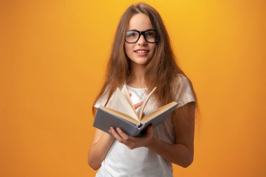 Gözlüklü zeki kız sarı arka planda kitap tutuyor.