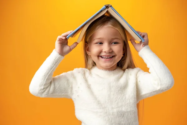 Маленькая девочка держит книгу на голове и смотрит в камеру на жёлтом фоне — стоковое фото