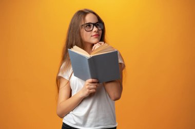 Smart teen girl in glasses holding book against yellow background