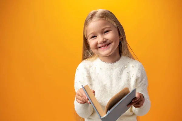 Little beautiful smiling girl holding book against yellow background — ストック写真