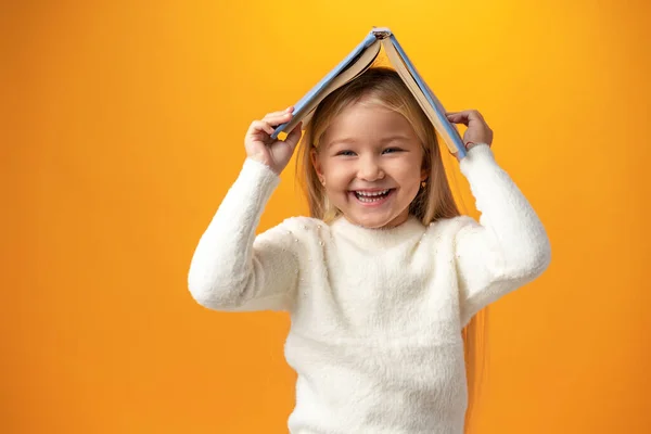 Маленькая девочка держит книгу на голове и смотрит в камеру на жёлтом фоне — стоковое фото