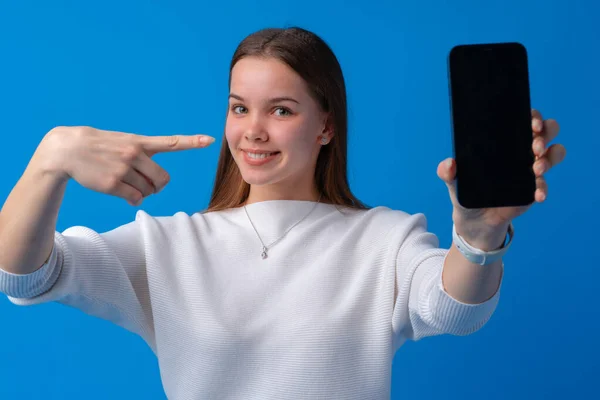 Portret uśmiechniętej kobiety pokazującej pusty ekran telefonu komórkowego na niebieskim tle — Zdjęcie stockowe