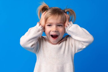 Elleri başının üstünde olan güzel küçük kız mavi arka planda seviniyor ve şok oluyor..