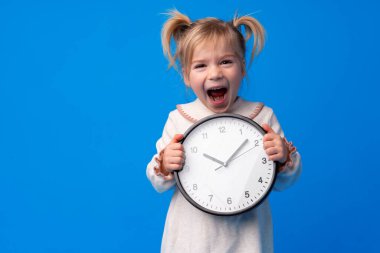 Güzel genç kız mavi arka planda duvar saatini tutuyor.