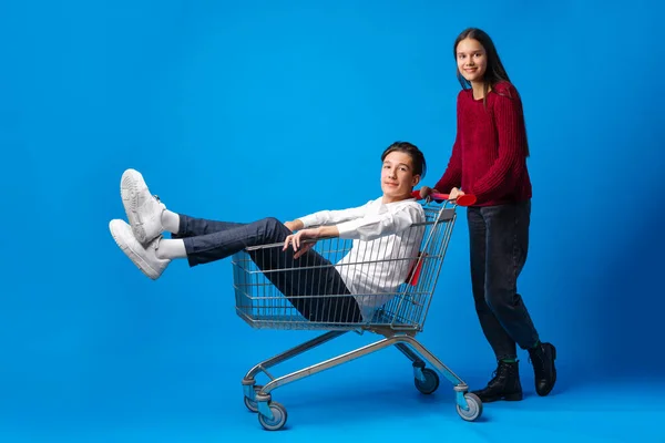 Adolescente empurrando seu namorado adolescente em um carrinho de compras contra fundo azul em estúdio — Fotografia de Stock