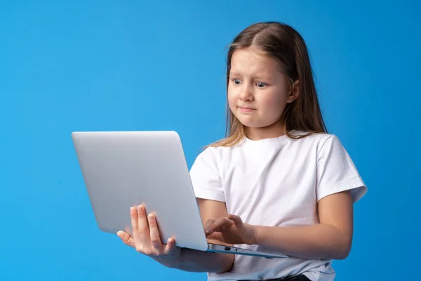 Retrato de una niña sosteniendo un ordenador portátil mientras está de pie sobre un fondo azul —  Fotos de Stock