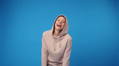 Bej kapüşonlu genç mutlu kadın mavi arka plana gülüyor.