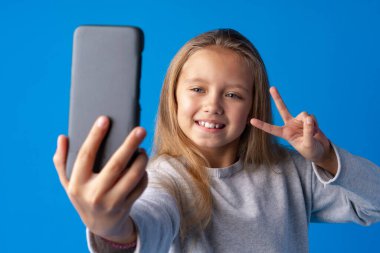 Heyecanlı küçük bir kızın portresi mavi arka planda izole edilmiş bir selfie çekiyor.