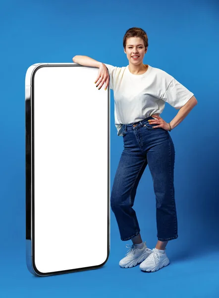 Glad ung kvinna lutar sig mot enorma mobiltelefon med tom vit skärm, för din annons, håna upp — Stockfoto