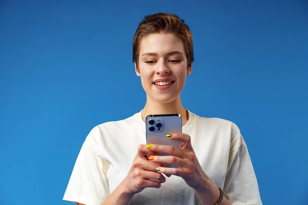 Fröhlich attraktive junge Frau hält Smartphone in der Hand, genießt Chat oder Online-Shopping vor blauem Hintergrund — Stockfoto