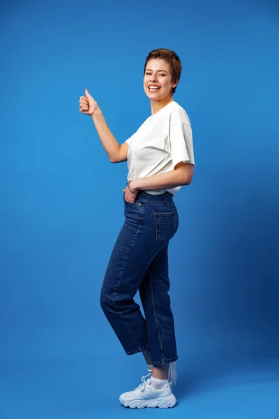 Joven feliz mujer alegre con el pelo corto mostrando el pulgar hacia arriba sobre fondo azul — Foto de Stock