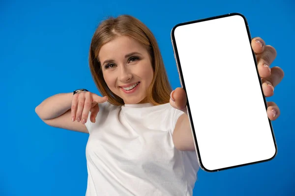 Mujer joven señala con el dedo la pantalla aislada del teléfono inteligente sobre fondo de color azul —  Fotos de Stock