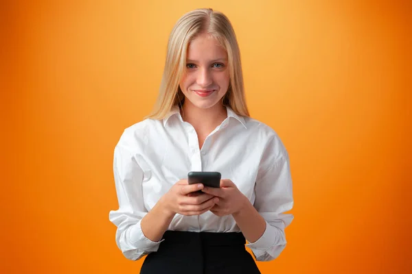 Menina adolescente bonito usando telefone celular com expressão positiva contra fundo laranja — Fotografia de Stock