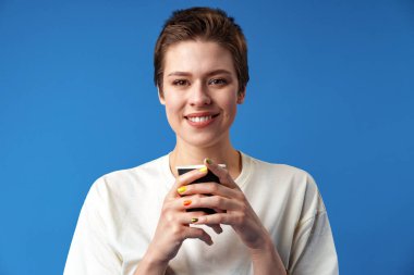 Güzel, gülümseyen kadın mavi arka planda kahve içiyor.