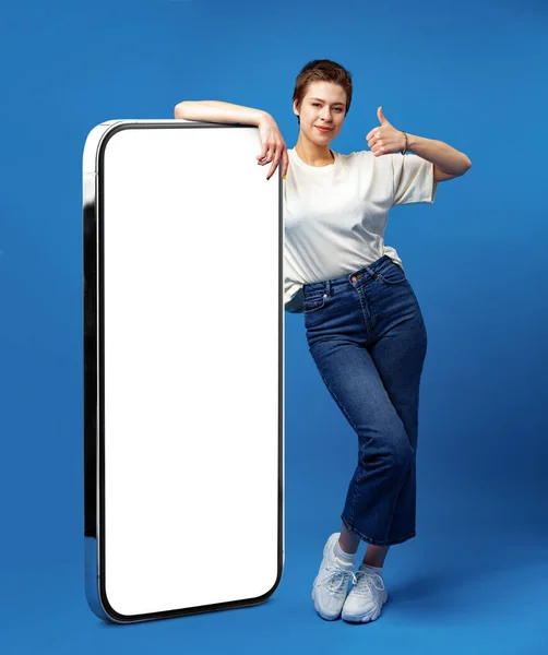 Joyeux jeune femme appuyé sur un énorme téléphone portable avec écran blanc vide, pour votre annonce, maquette — Photo