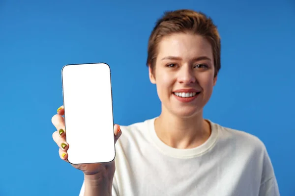 Portrait d'une jeune femme montrant un téléphone portable à écran vierge debout sur fond bleu — Photo