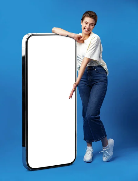 幸せな若い女性は空の白い画面で巨大な携帯電話にもたれて、あなたの広告のために、モックアップ — ストック写真
