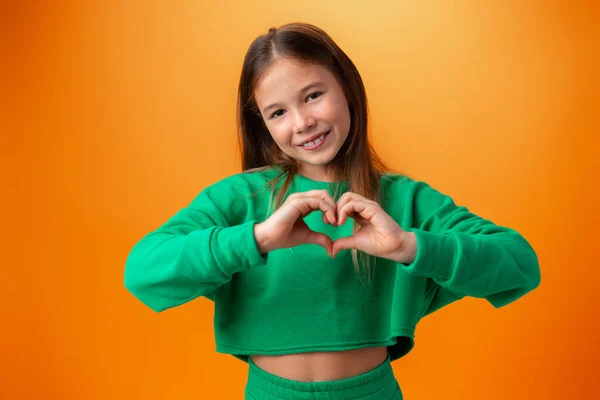 Ragazza adolescente che mostra il cuore alla macchina fotografica mentre in piedi su sfondo arancione — Foto Stock