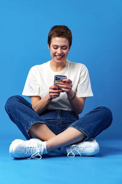 Vidám vonzó fiatal nő gazdaság smartphone, élvezi a csevegés vagy online vásárlás a kék háttér — Stock Fotó